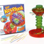 The game KerPluck. Shown here are the game box and the game itself. A plastic tower, some straws and marbles. The aim of the game is to pull out the straws and not allow the marbles to fall. The winner is the player with the least marbles at the end.