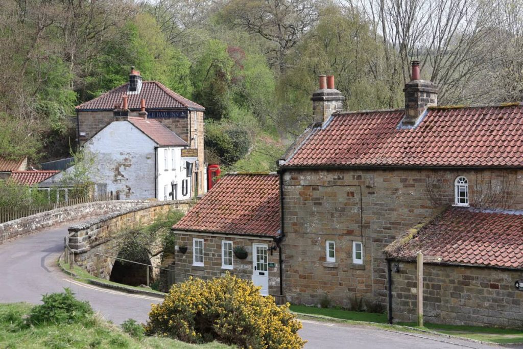 A Yorkshire village with a few houses and a pub in the background. There is a stone bridge crossing a river.