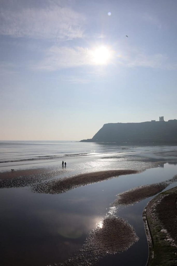 Two people walking on a deserted beach with a castle in the background. The morning sun is in the sky.