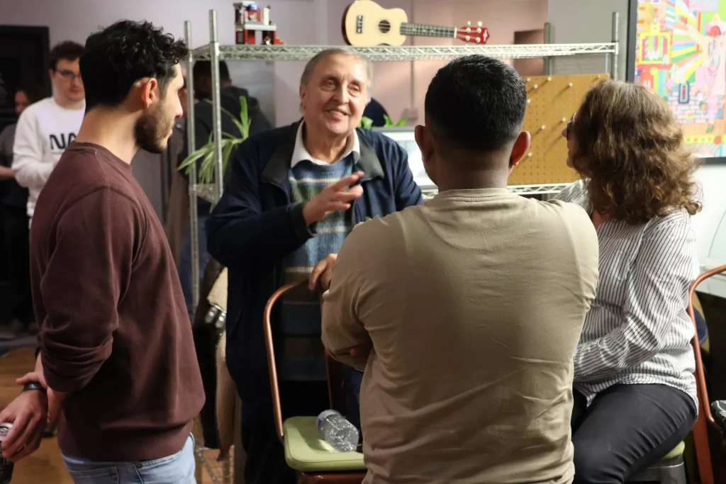 A group of four people engaged in conversation at a networking event, with one person gesturing while speaking. Background features shelves with various items.