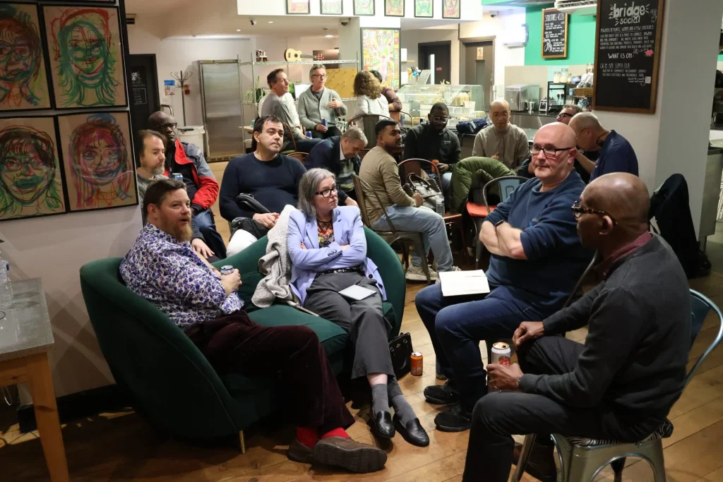 A group of people seated in a cozy café setting, engaged in conversation. Some attendees are taking notes, while others appear to be listening intently to the discussion.
