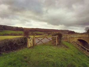 A wooden gate opening to a rural landscape, with rolling hills and a stone bridge in the background under cloudy skies.