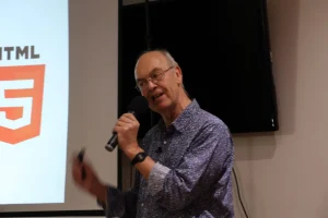 A person speaking into a microphone during a presentation with a large HTML5 logo displayed in the background.