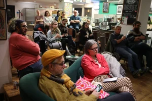 A group of people seated in a cozy café setting, attentively listening to a presentation or discussion. Various individuals display a range of expressions, with some looking at their phones and others engaging with the speaker.