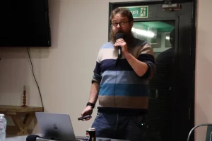 A person with a beard wearing a striped sweater stands at a podium, holding a microphone and gesturing while speaking to an audience.