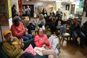 A lively audience clapping in a cozy indoor setting, with seated attendees wearing colorful clothing and displaying enthusiasm, while others stand in the background.