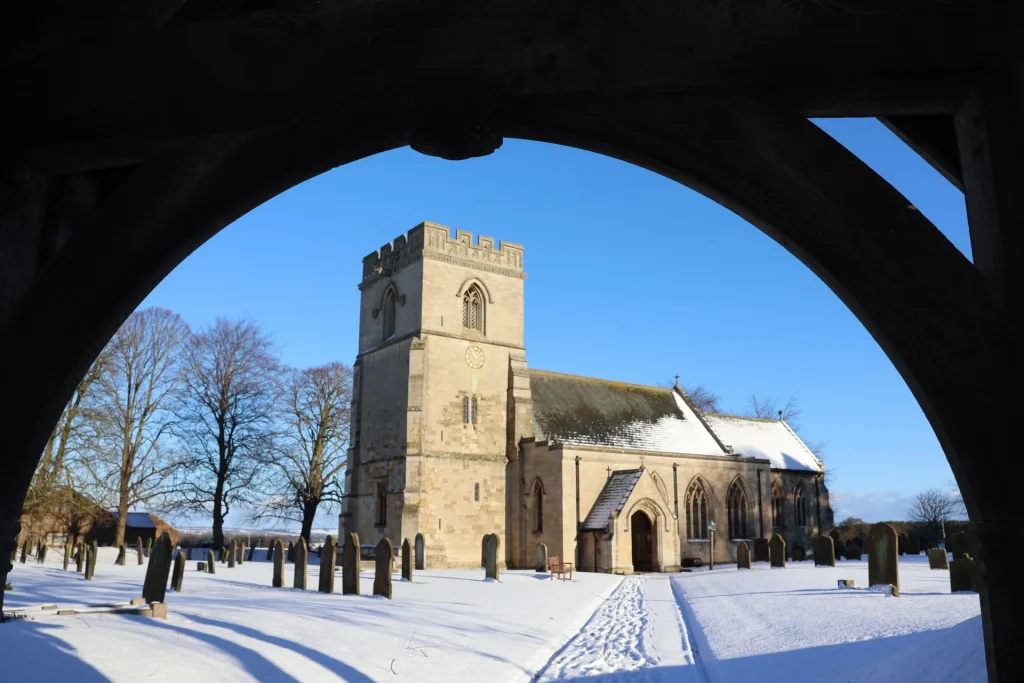 A snow-covered church surrounded by trees and gravestones, viewed through a wooden archway.