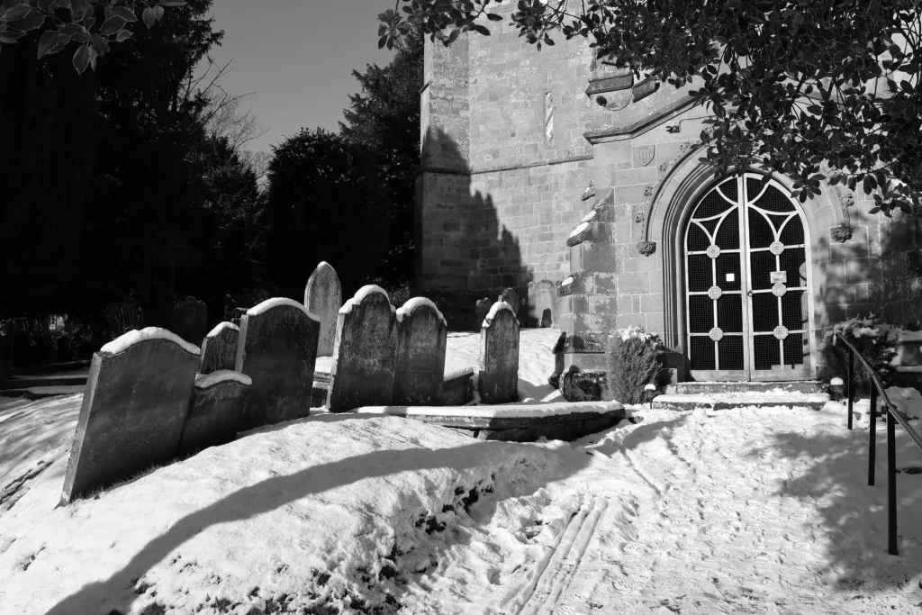 Black and white image of a snowy graveyard with several gravestones partially covered in snow, in front of a stone church entrance.