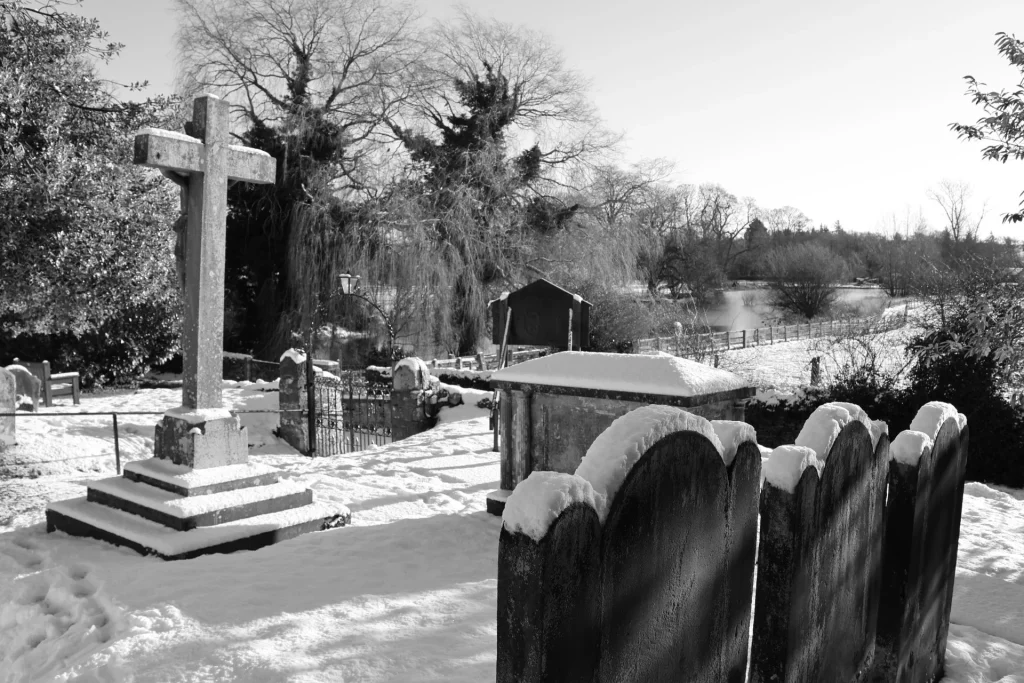 A snowy cemetery scene featuring grave markers and a cross, with a peaceful landscape in the background.