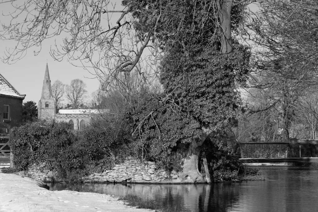A monochrome photograph of a snowy landscape featuring a large tree covered in ivy, a church with a tall spire in the background, and a calm river in the foreground.