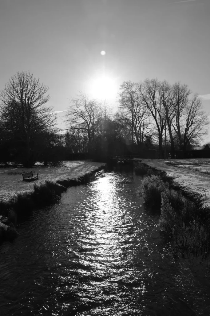 Black and white photograph of a river reflecting sunlight, with a bench on the snowy bank and bare trees silhouetted against the sky.