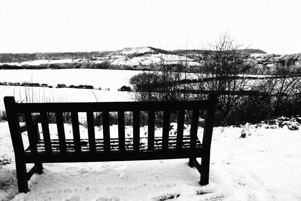 A black and white photograph of a wooden bench in a snowy landscape, overlooking hills blanketed in snow.
