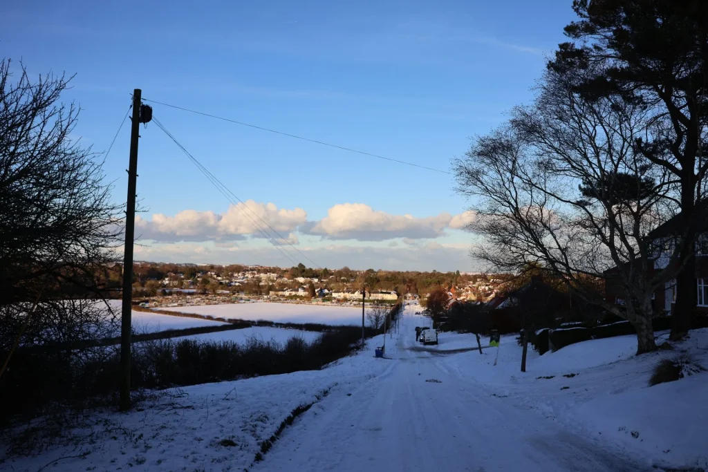 A snowy landscape featuring a cleared road, power lines, and trees on either side, with a view of houses in the distance under a blue sky with white clouds.