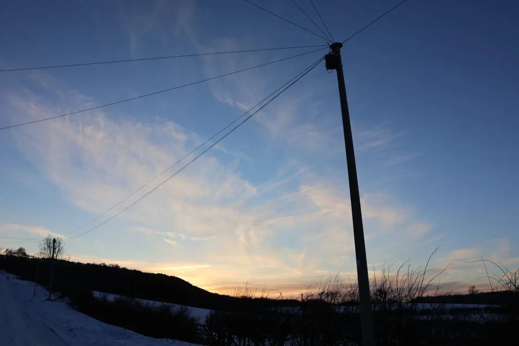 A winter sunset with soft clouds in the sky, silhouetted by a power pole and trees, amidst a snowy landscape.