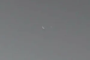 A close-up image of the crescent moon Phobos, Mars' moon, next to a small dot representing Earth, taken by NASA's Curiosity rover on the surface of Mars.