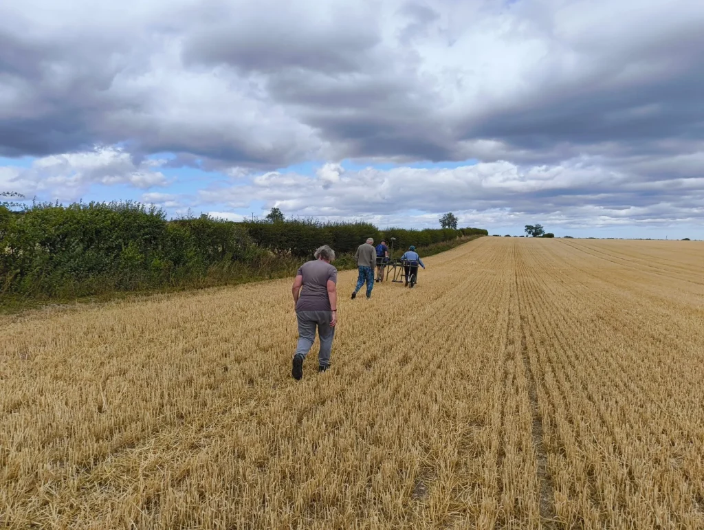 Four people walking through a harvested wheat field under a partly cloudy sky, with hedgerows in the background.