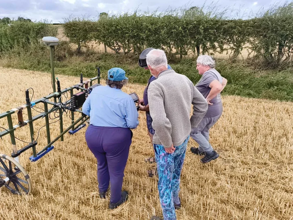 Four people interacting around archaeological equipment in a field, examining a digital display.